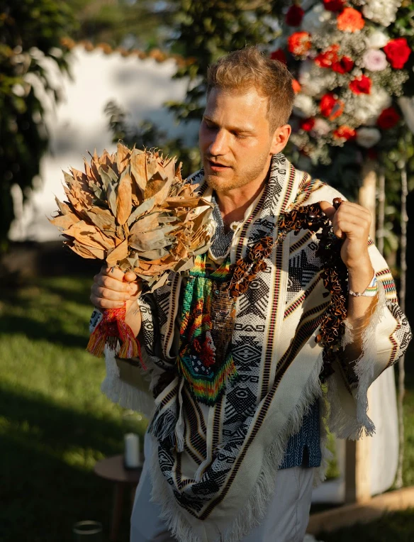 Oliver Glozik in a woven poncho, holding a bundle of dried leaves and a seed necklace at an outdoor ceremony