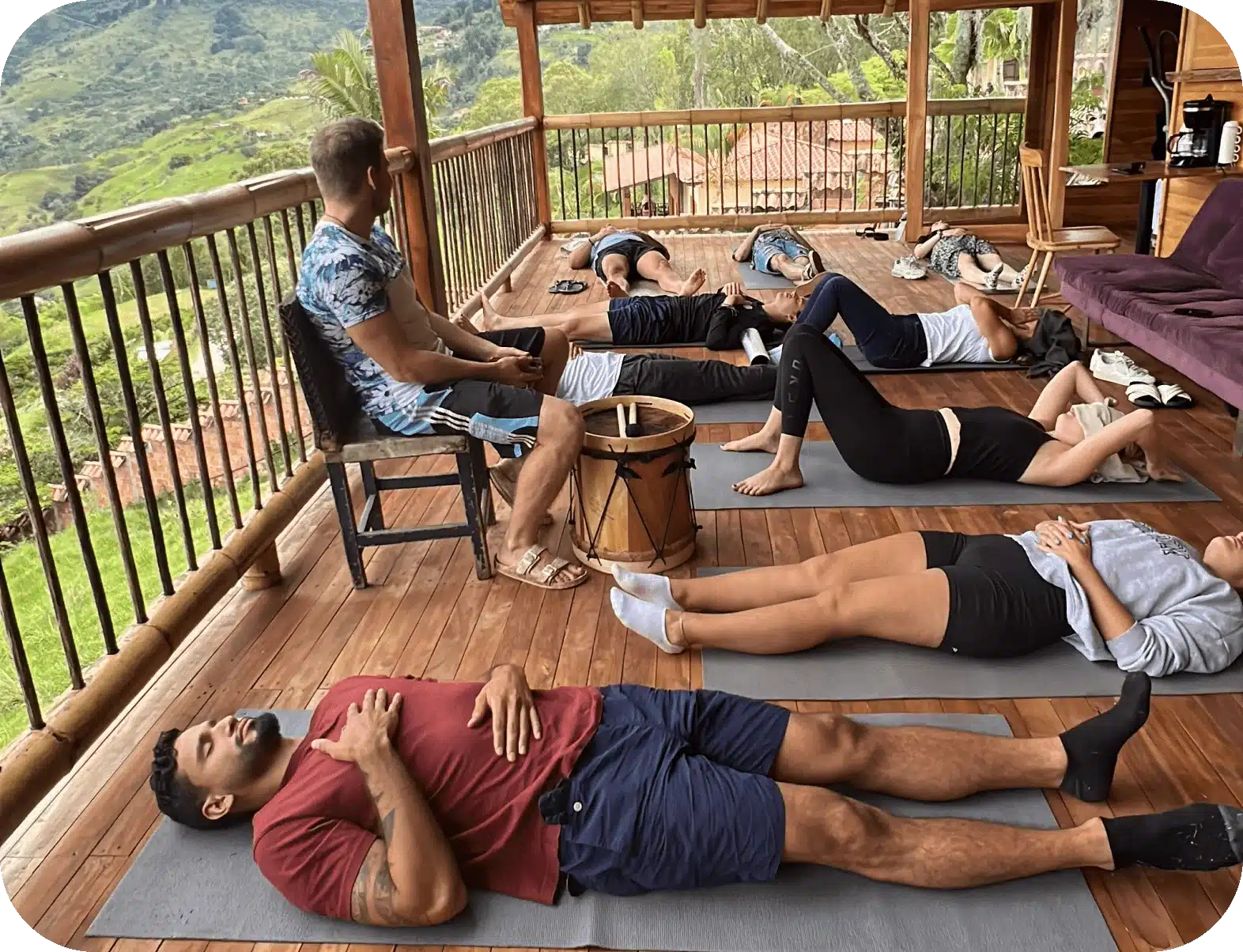 Oliver sitting beside a drum on a wooden balcony above green mountains, guiding a group lying on mats through breathwork