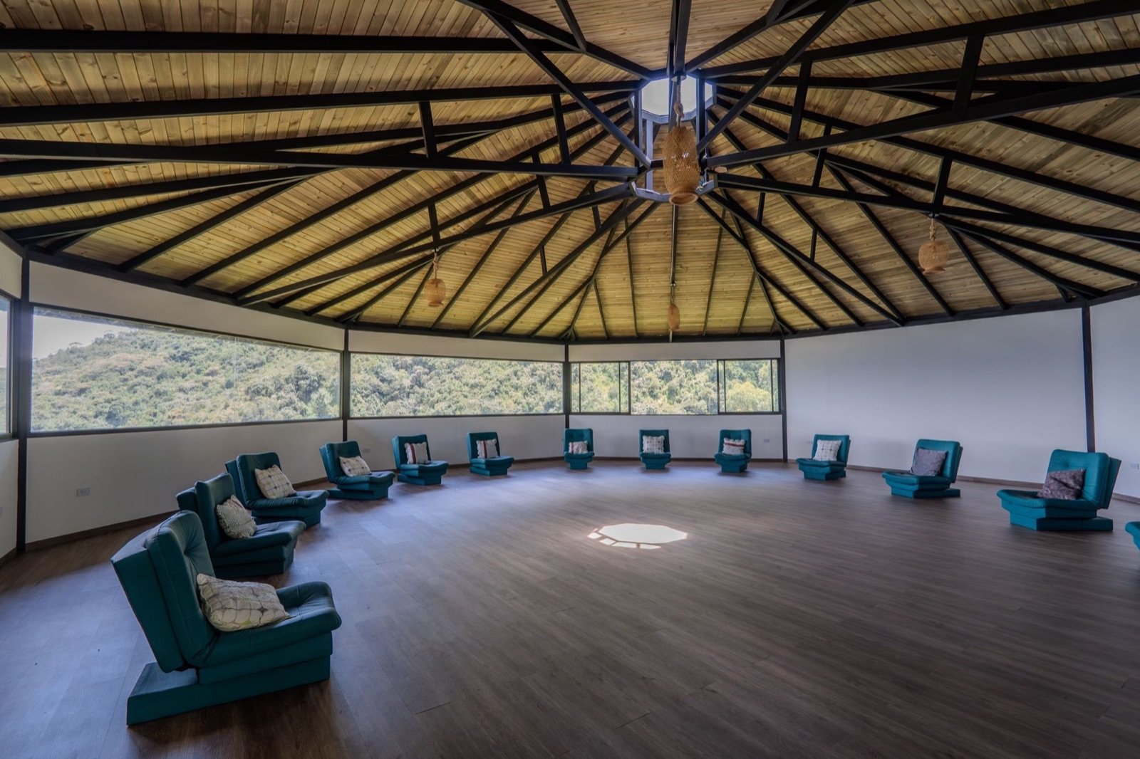 A round group room with a wooden roof, a circle of teal armchairs and wide windows onto forested hills